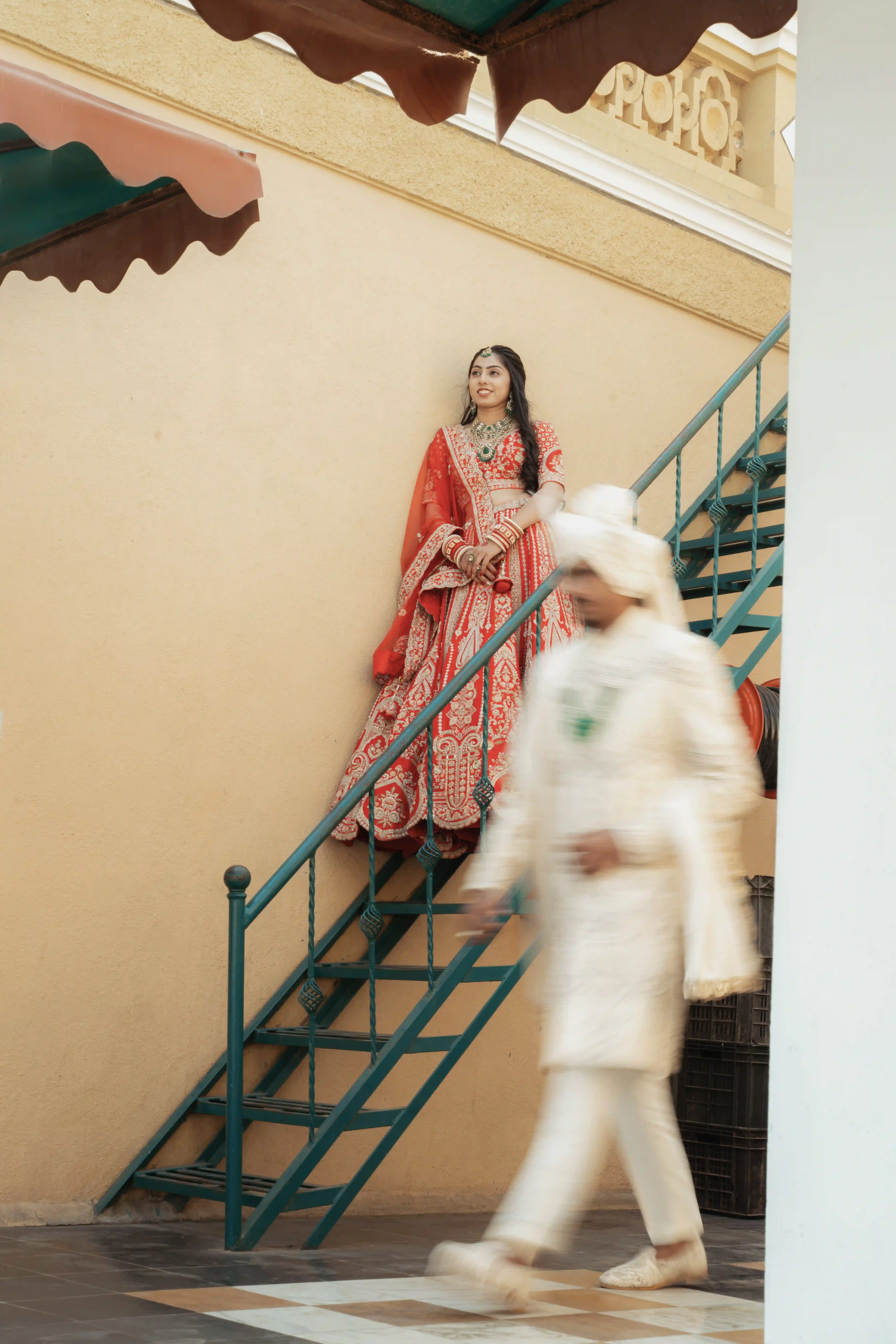 Bride in red bridal lehenga standing on staircase with groom walking past